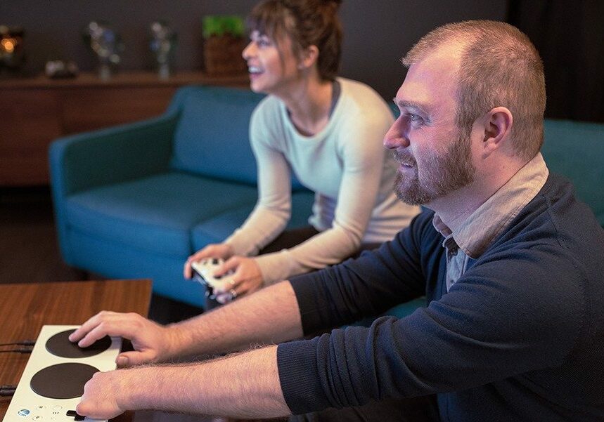 A man and woman playing video games sitting on a couch smiling. The man is using the microsoft accessible tool