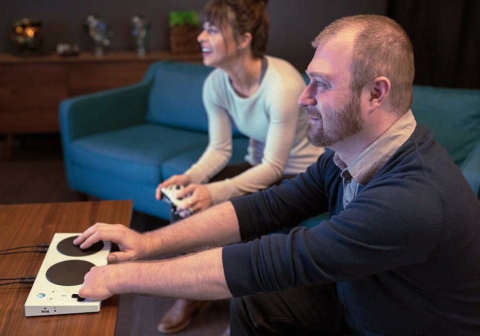 A man and woman playing video games sitting on a couch smiling. The man is using the microsoft accessible tool